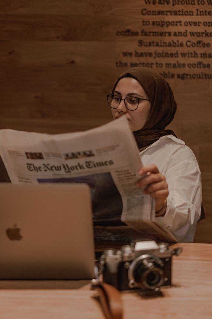 creative-03 Muslim woman in hijab reading a newspaper at a café, laptop and camera on table.