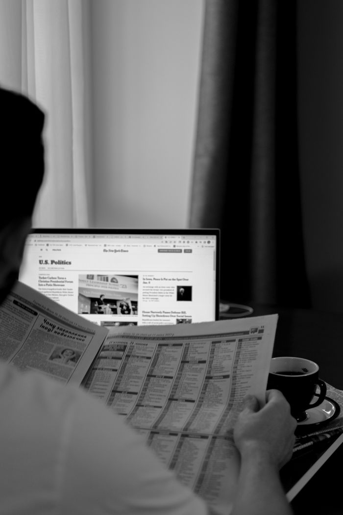 creative-02 Black and white photo of a man reading a newspaper with a laptop in the background.