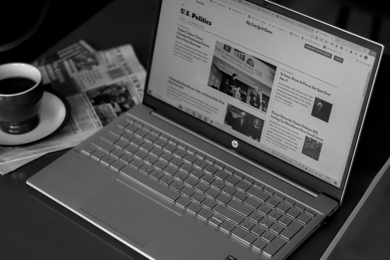Services-02 Black and white image of a laptop displaying news articles, accompanied by a cup of coffee and newspapers.