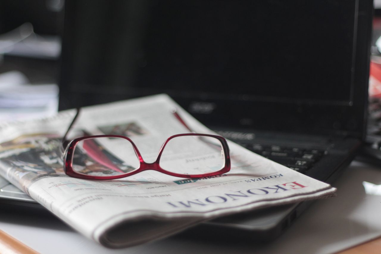 mobile-01 Close-up of red eyeglasses on a newspaper with a laptop in the background, suggesting reading or work.